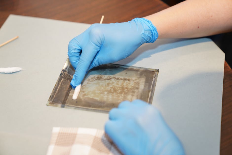 Hands in gloves carefully restoring vintage artwork on glass using cleaning tools.